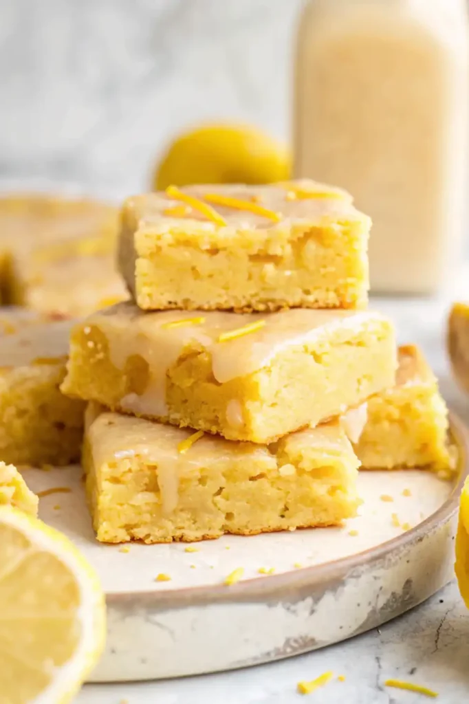 A stack of lemon brownies with a glossy glaze and lemon zest on a ceramic plate.