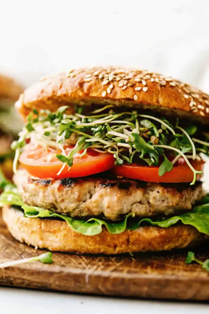 A stacked turkey burger on a sesame bun with fresh greens, sliced tomatoes, and sprouts on a rustic wooden board.