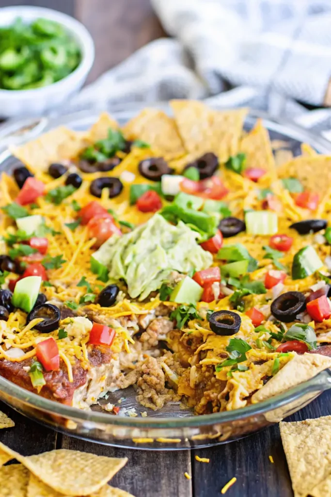 A layered taco dip topped with guacamole, black olives, and chopped tomatoes, served with tortilla chips.